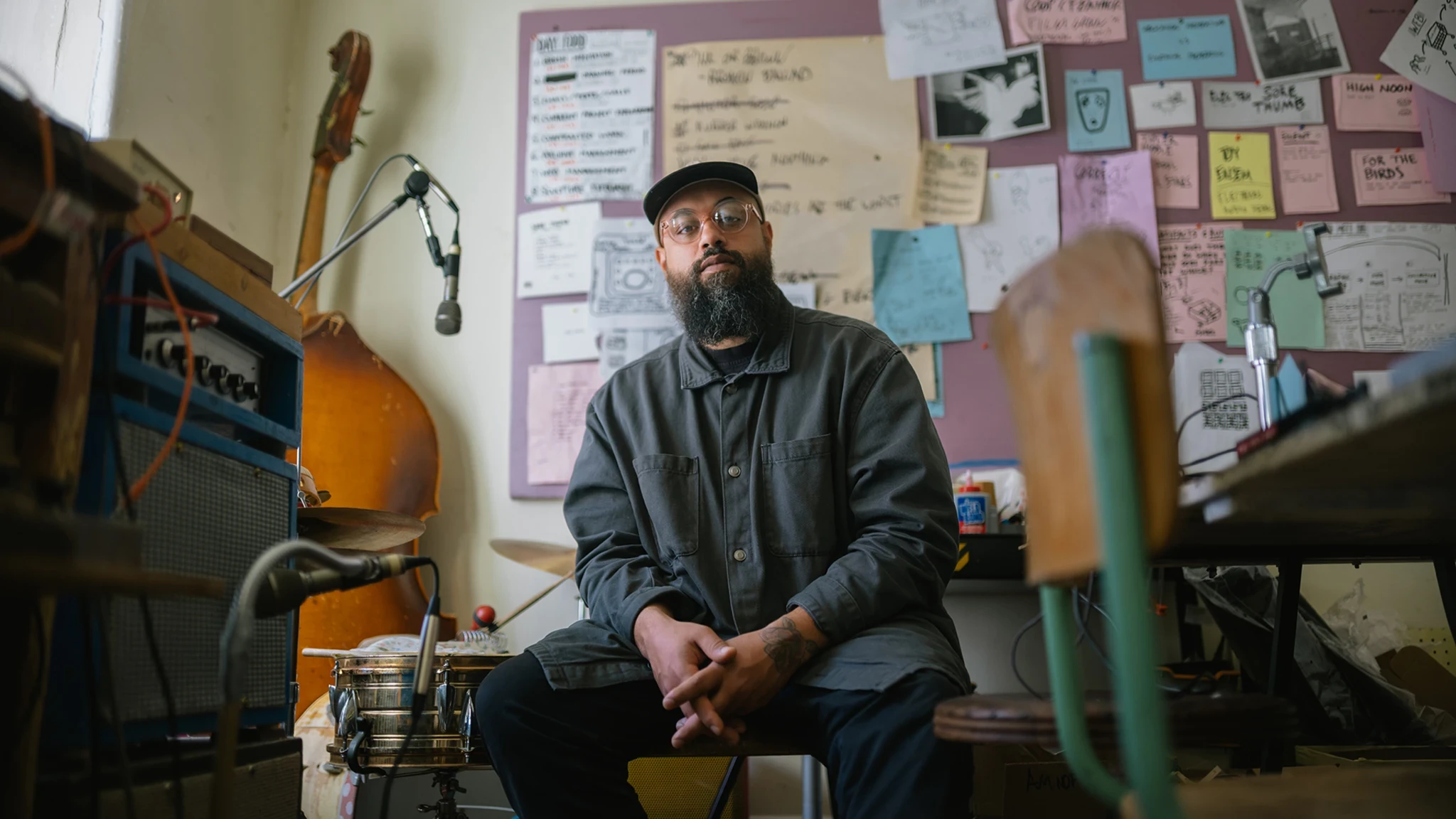 A man with a beard wearing glasses, a hat, and a blue shirt sits in a room surrounded by instruments and poster art.