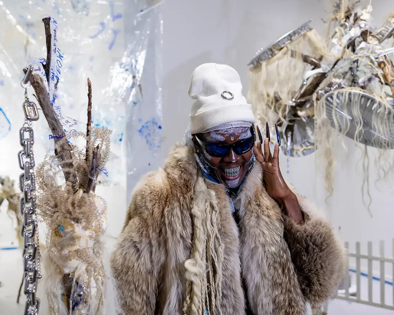 Vitche-Boul Ra wears a fur coat, white hat, sunglasses, a bejeweled grill, and long black fingernails.