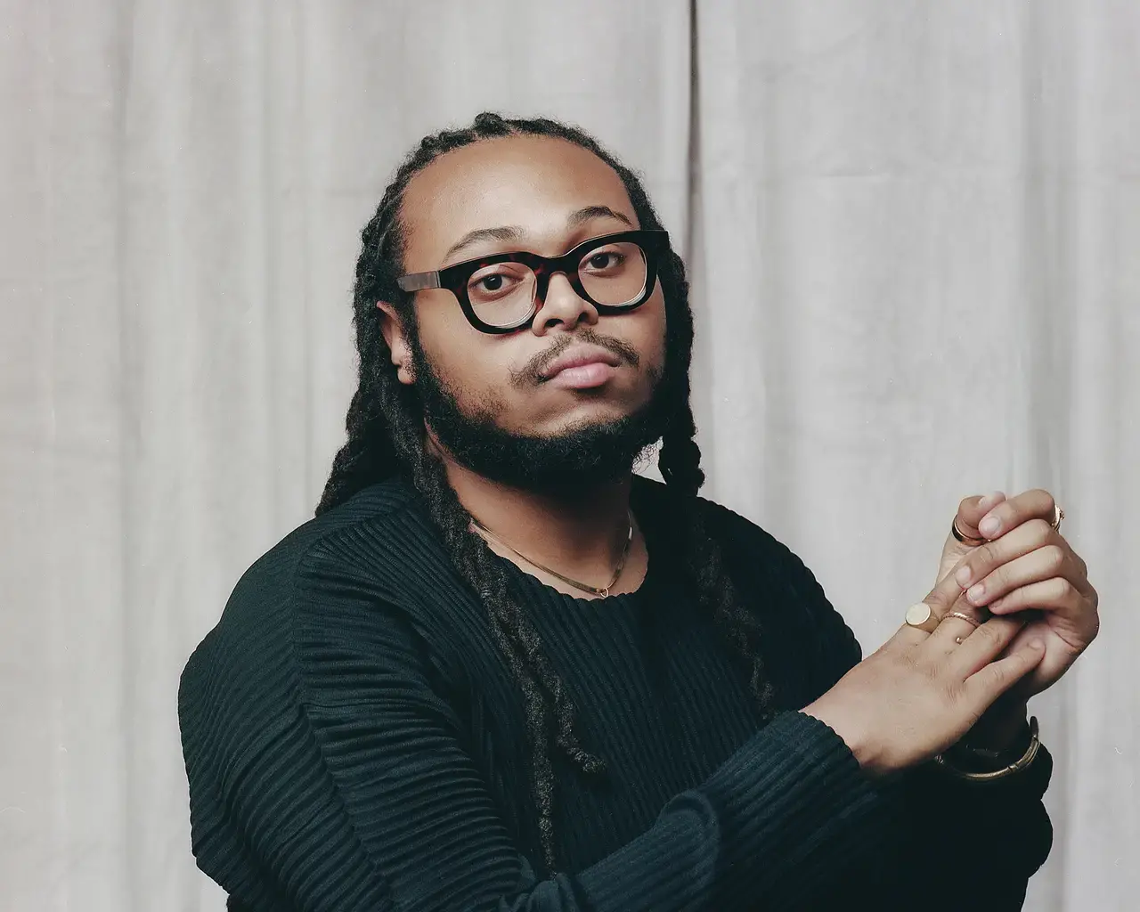 A portrait shot of saxaphone player Immanuel Wilkins wearing a black ribbed shirt, dark rimmed glasses, and long black twists.