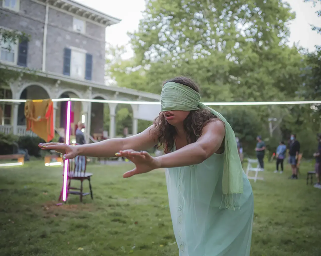 A performer wears a seafoam colored blindfold and gauzy blue dress. Their arms are extended and searching.