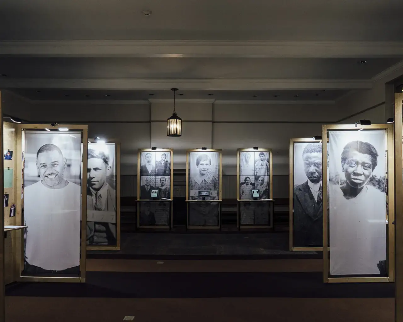 Six, larger than life-sized portraits of men and women line either side of a carpeted room. The images are printed on fabric and hung from wooden frames. In the back of the room, three additional wooden frames; the center frame contains a portrait, and the frames on either side of the portrait contain a grid of six images each and abstracted archival text. All but two of the portraits in the room are archival.