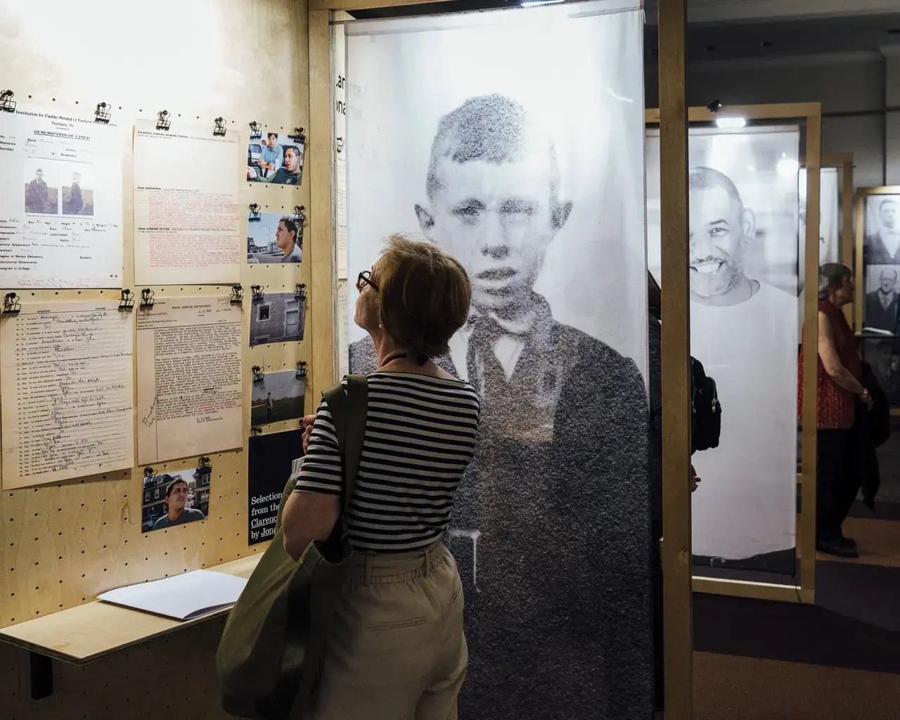 A middle-aged white woman with short red hair stands in front of pegboard display panel, with an adjacent archival portrait of a young man wearing a suit and tie. She is reading the material displayed, which includes archival documents and images, modern day photos and directional text. A Braille book sits on a wooden shelf attached to the display panel.