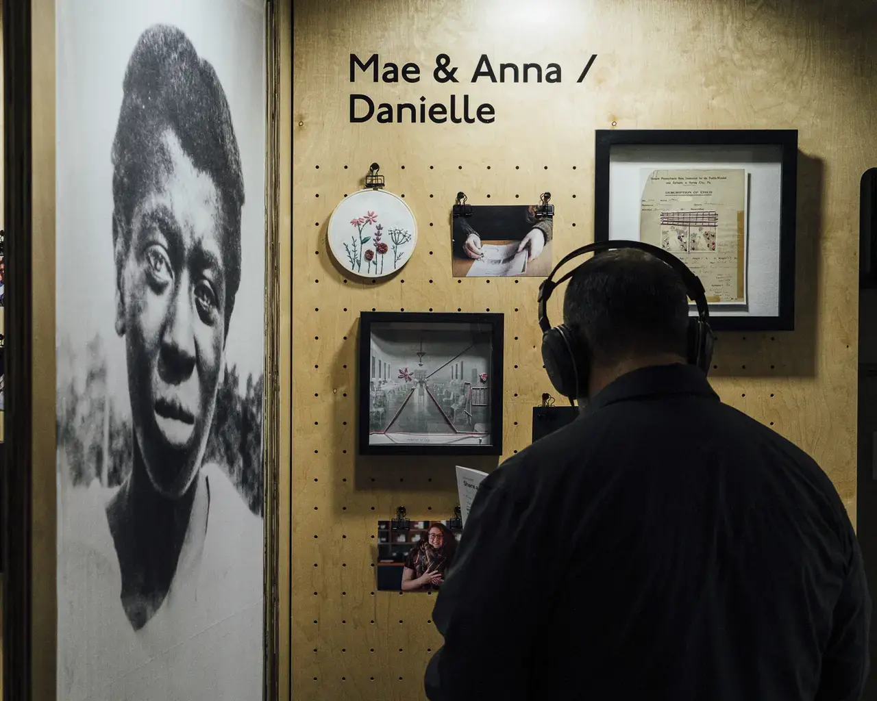 A man wearing headphones stand in front of a wooden display that features framed embroidery, archival imagery and contemporary photos. The words Mae & Anna / Danielle appear on the display. Adjacent to the display is a larger than life-sized archival portrait of a middle-aged Black woman.