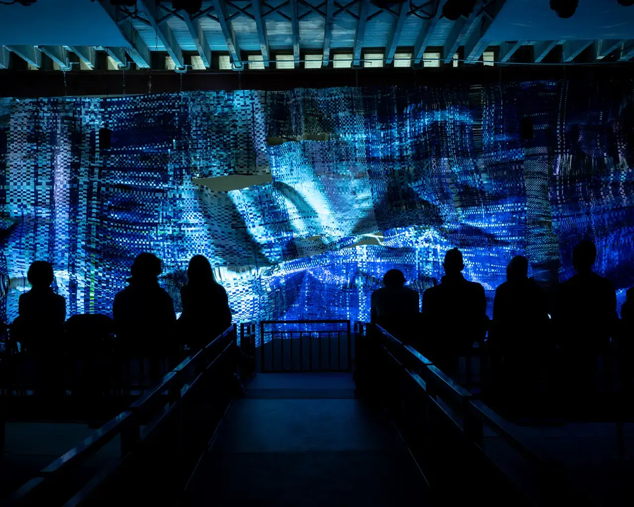 Audience members sit in a dark viewing space in front of a bright blue cyanotype weaving.