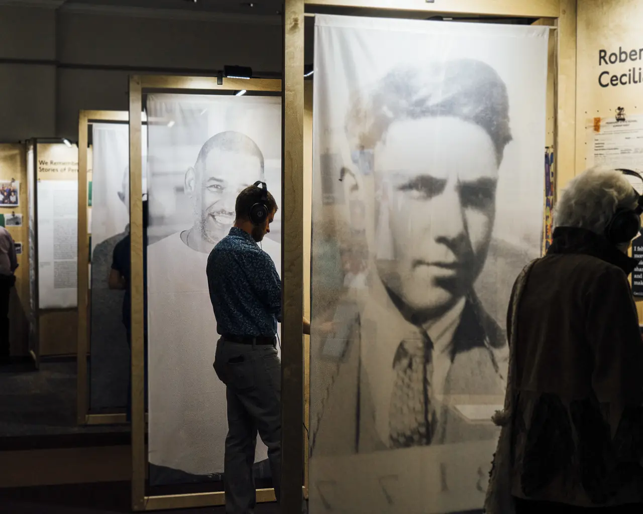 Image description: A row of large wooden display panels featuring three, larger-than-life portraits of men, tow archival and one modern. A woman stands at the first display, and a man stands at the second display. Both are wearing headphones. A third man stands at a display in the back of the room.