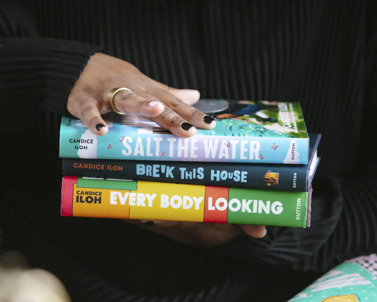 A pair of brown-skinned hands with black nail polish hold a stack of three coloroful books, their spines facing the camera. The holder is wearking a black sweater.