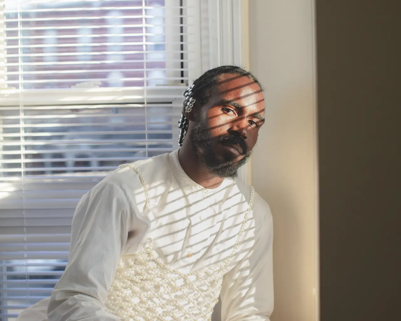 A portrait of filmmaker Vernon Jordan. He sits leaning against a window with the shadow of the blinds cascading across his face and body.