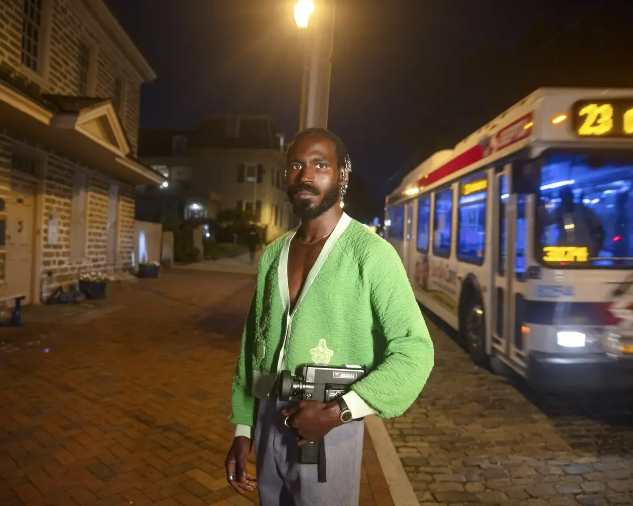 A portrait of filmmaker Vernon Jordan. He stands outside at night holding his video camera. He is a Black man with facial hair wearing a green v-neck cardigan. A SEPTA bus is visible come down the street behind him.