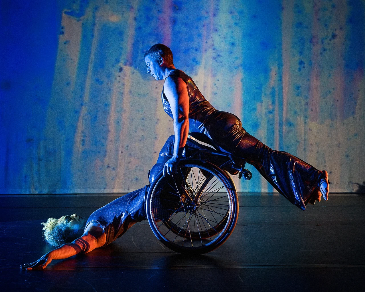 A moment from Kinetic Light’s The Next TiMes. Laurel Lawson holds their body in a diagonal line as they balance atop Alice Sheppard’s wheels and shins. Alice supports from below as they lay back, arms and torso on the ground, hips lifted. Alice is a multiracial Black woman with short curly hair, Laurel is a white person with very short, cropped hair; they both wear shimmery sleeveless bodysuits. Behind them, a projection of soft blues, purples, and greens looks like rain on a windowpane.