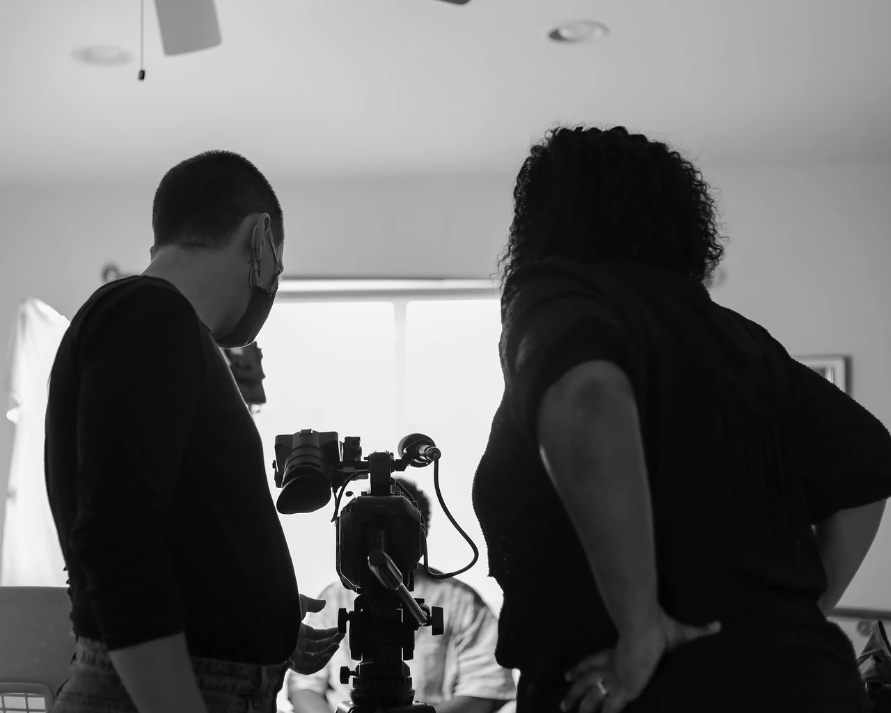 A black and white photo of director Jos Duncan Asé and cinematographer Marie Hinson standing in front of a digital video camera mounted to a tripod, on a location shoot in a living room.
