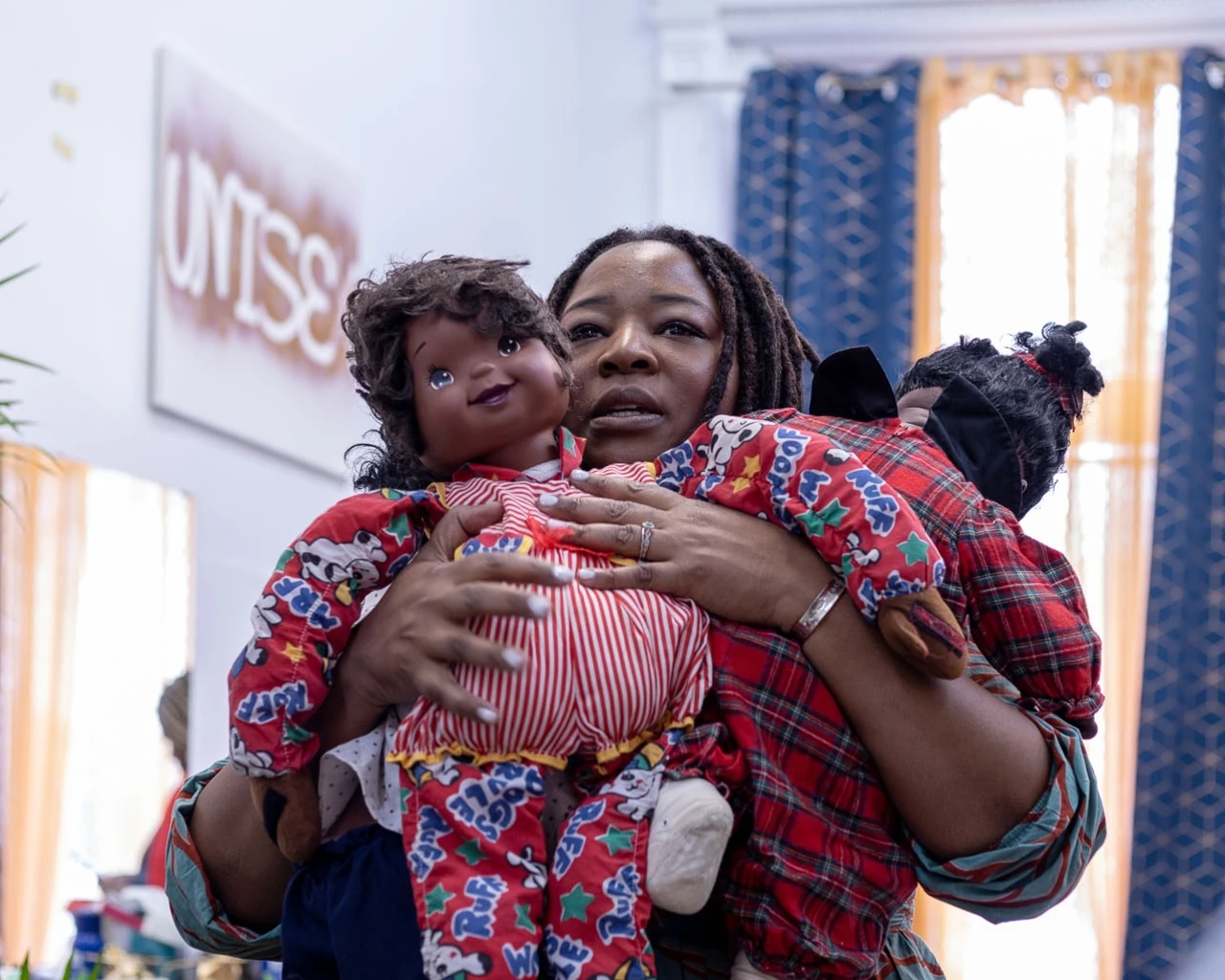 Jos Duncan Asé holding dolls during a location shoot in a living room, in front of a window with blue and yellow curtains.