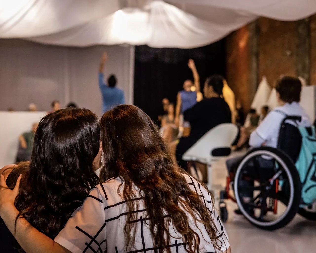 Rhythm Bath, 2023. &nbsp;Two women, each with long, wavy, dark hair, sit on the floor of a performance space. They are pictured from the back, heads close together, one woman’s arm wrapped around the other woman’s shoulder. In front of them, a dance performance takes place. Audience members stand or sit in chairs; one audience member uses a wheelchair.&nbsp;Photo by Ashley Smith, Wide-Eyed Studios.