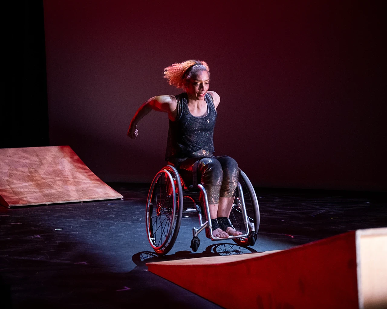 A moment from Kinetic Light’s Under Momentum. Alice Sheppard zooms up a wooden ramp in her chair, curved arms extended behind her and hair flying from the momentum of the movement. Alice is a multiracial Black woman with coffee-colored skin and short curly hair; she wears a shimmery sleeveless costume. Another ramp sits expectantly behind her. Photo by Christopher Duggan/Lincoln Center.&nbsp;