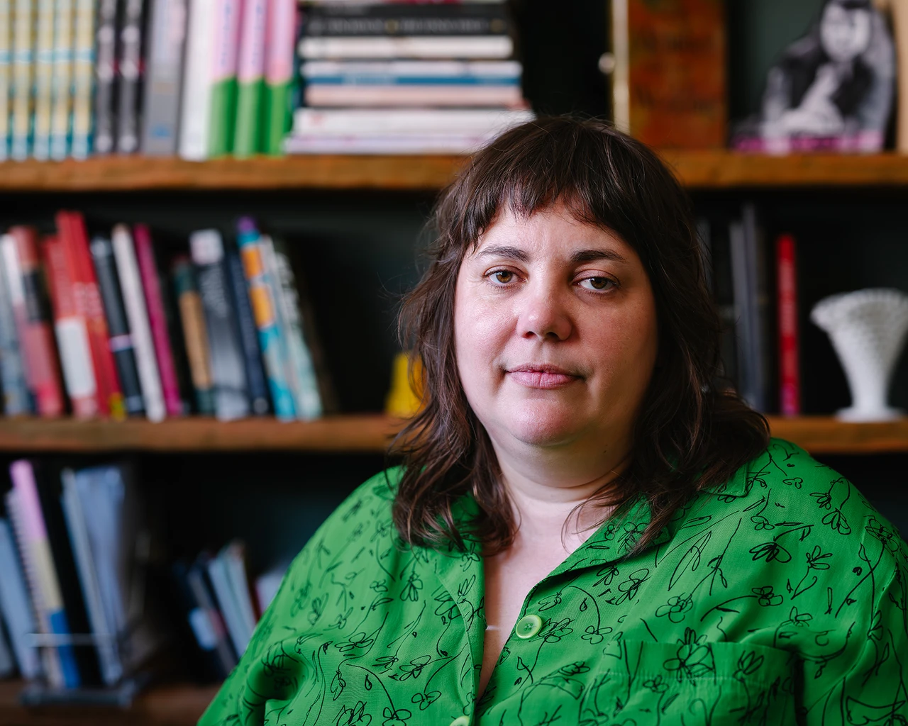 A white woman in a green shirt sits in front of a bookshelf.