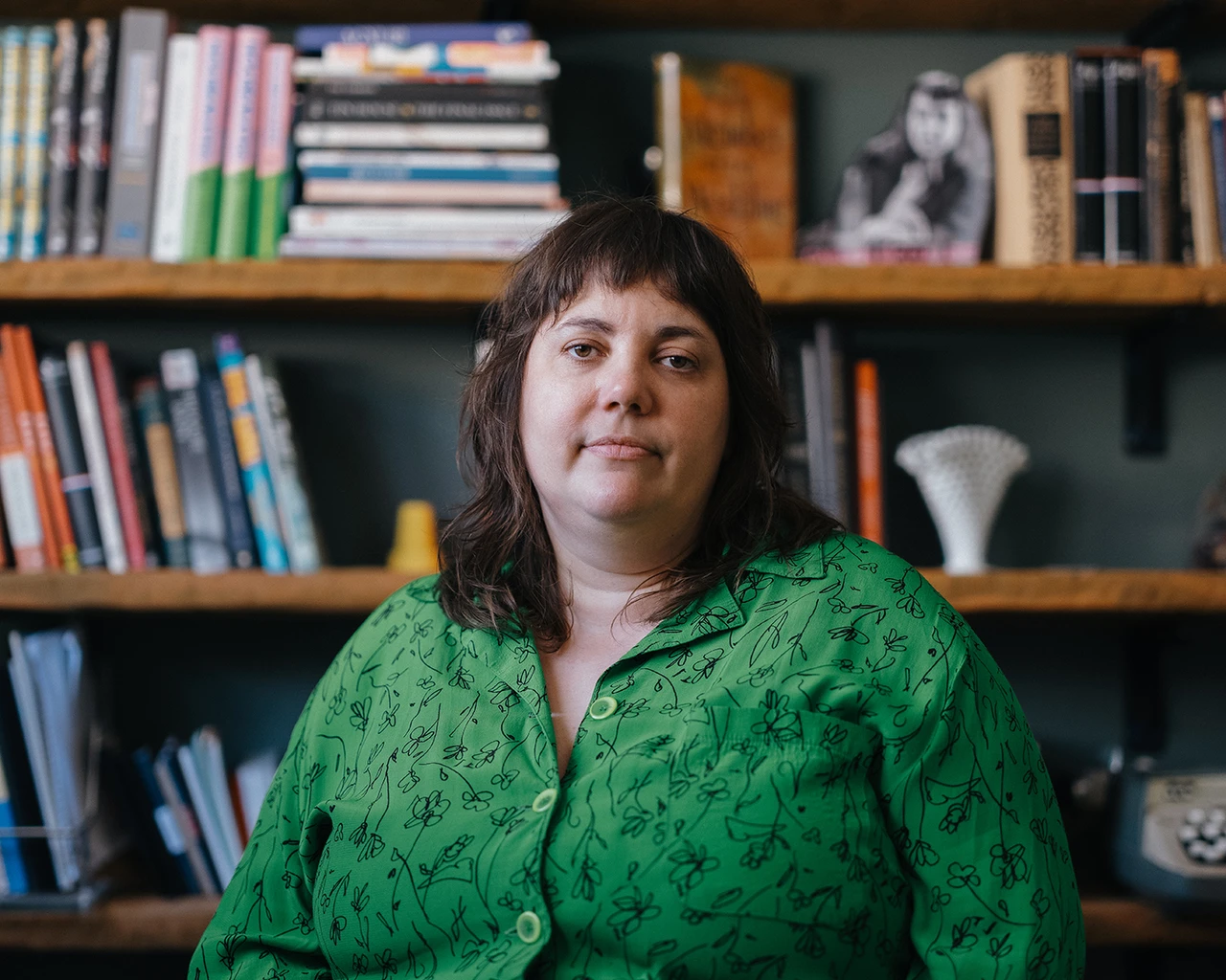 A white woman in a green shirt sits in front of a bookshelf.