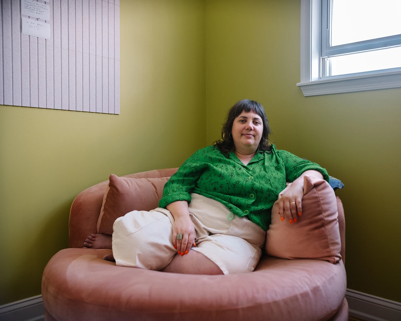 A white woman in a green shirt and white pants sits on a pink cushioned chairl