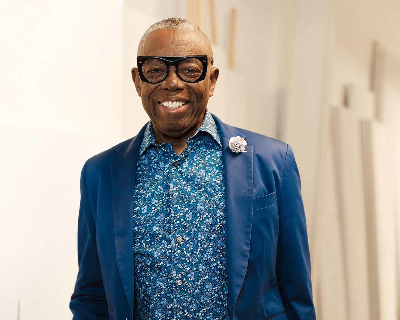 A Black man in a blue blazer and paisley shirt with glasses stands in front of a canvas.