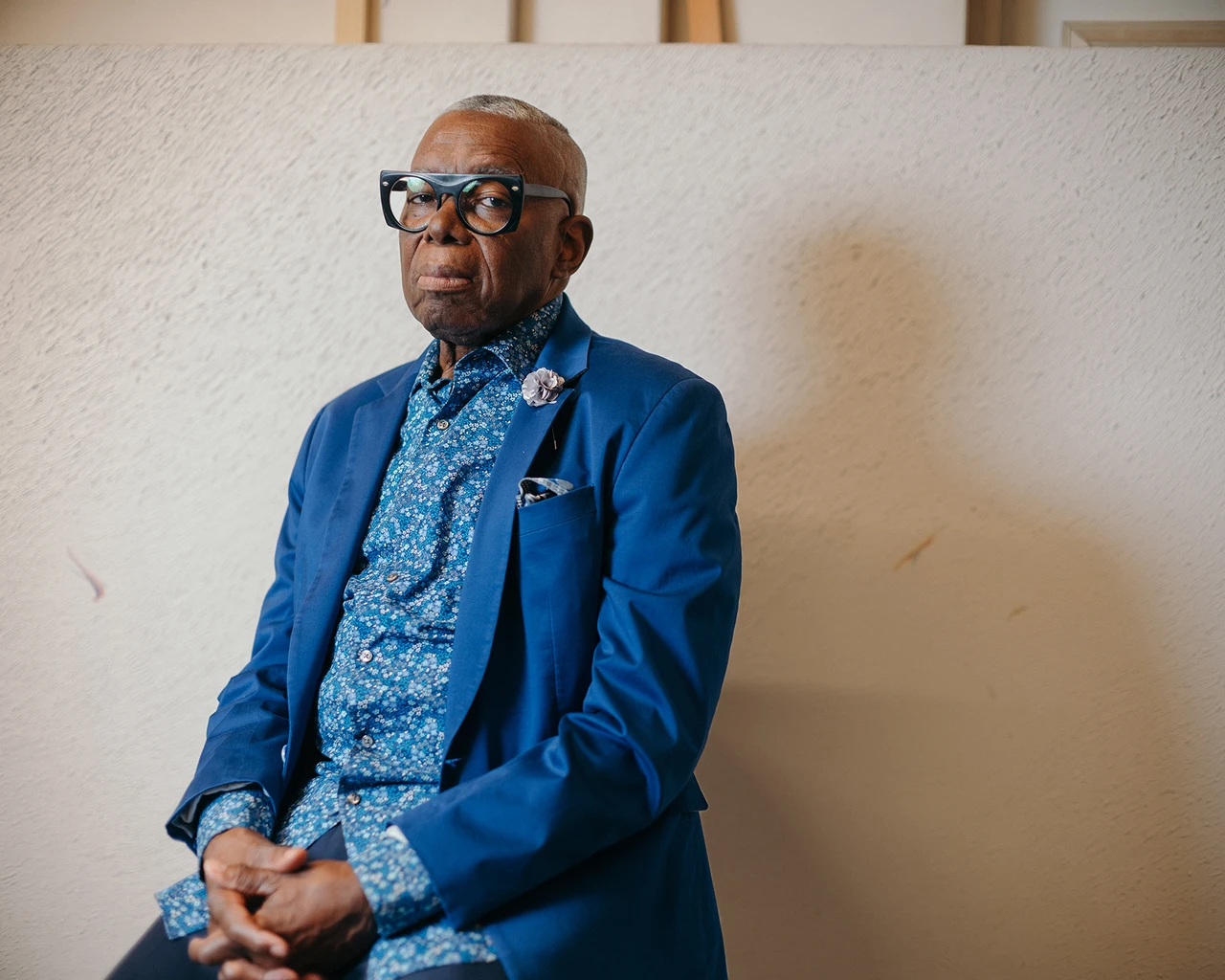 A Black man in a blue blazer and paisley shirt with glasses sits in front of a canvas.