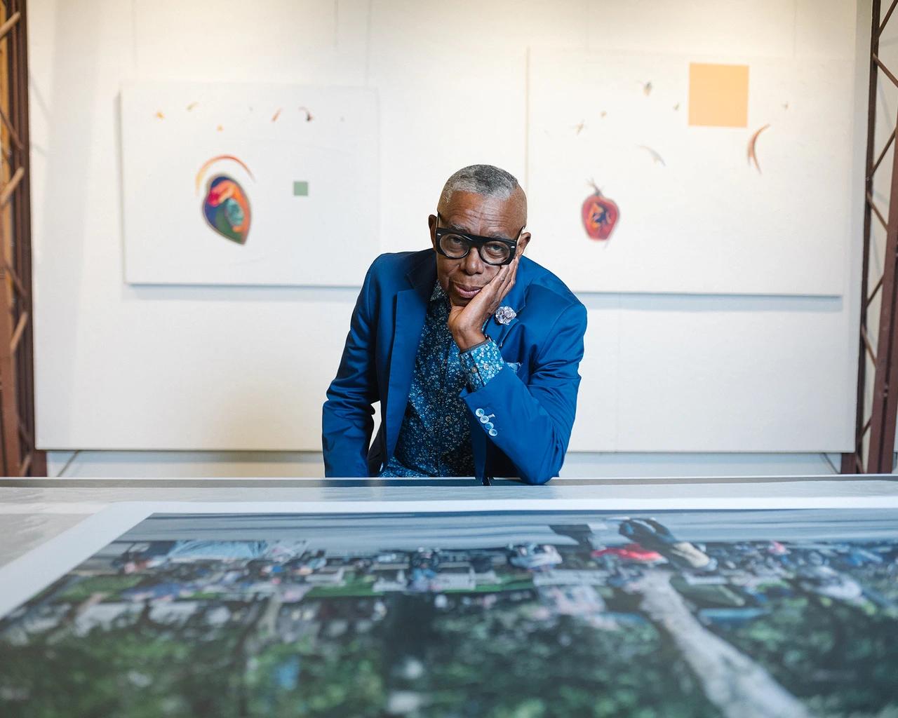 A Black man in a blue blazer and paisley shirt with glasses sits in front of a canvas.