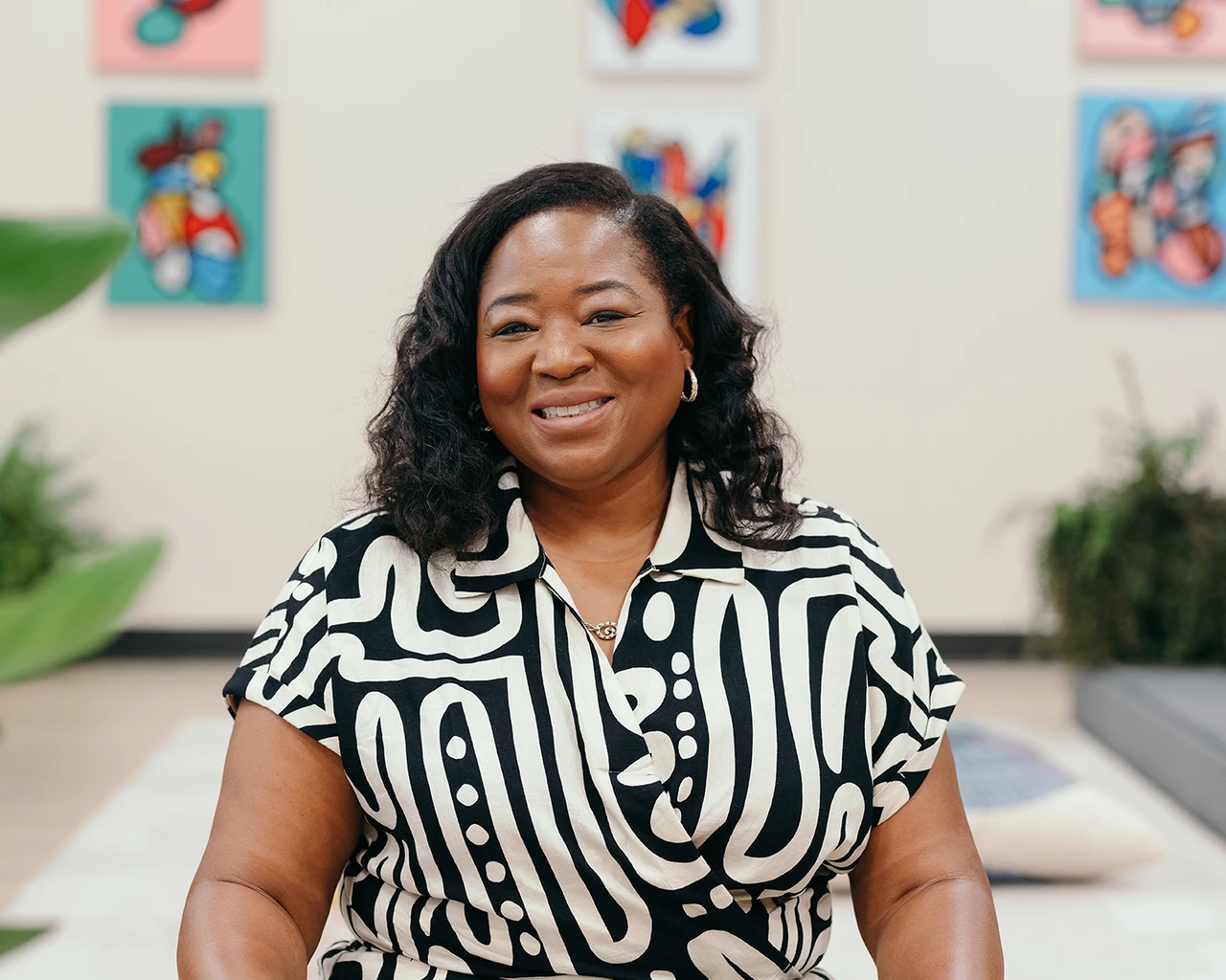A Black woman in a black and white patterened shirt sits in front of art prints.
