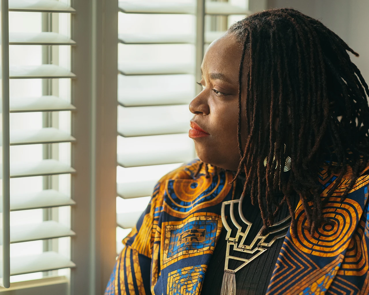 A Black woman in a yellow, blue, and maroon blazer sits at a window.