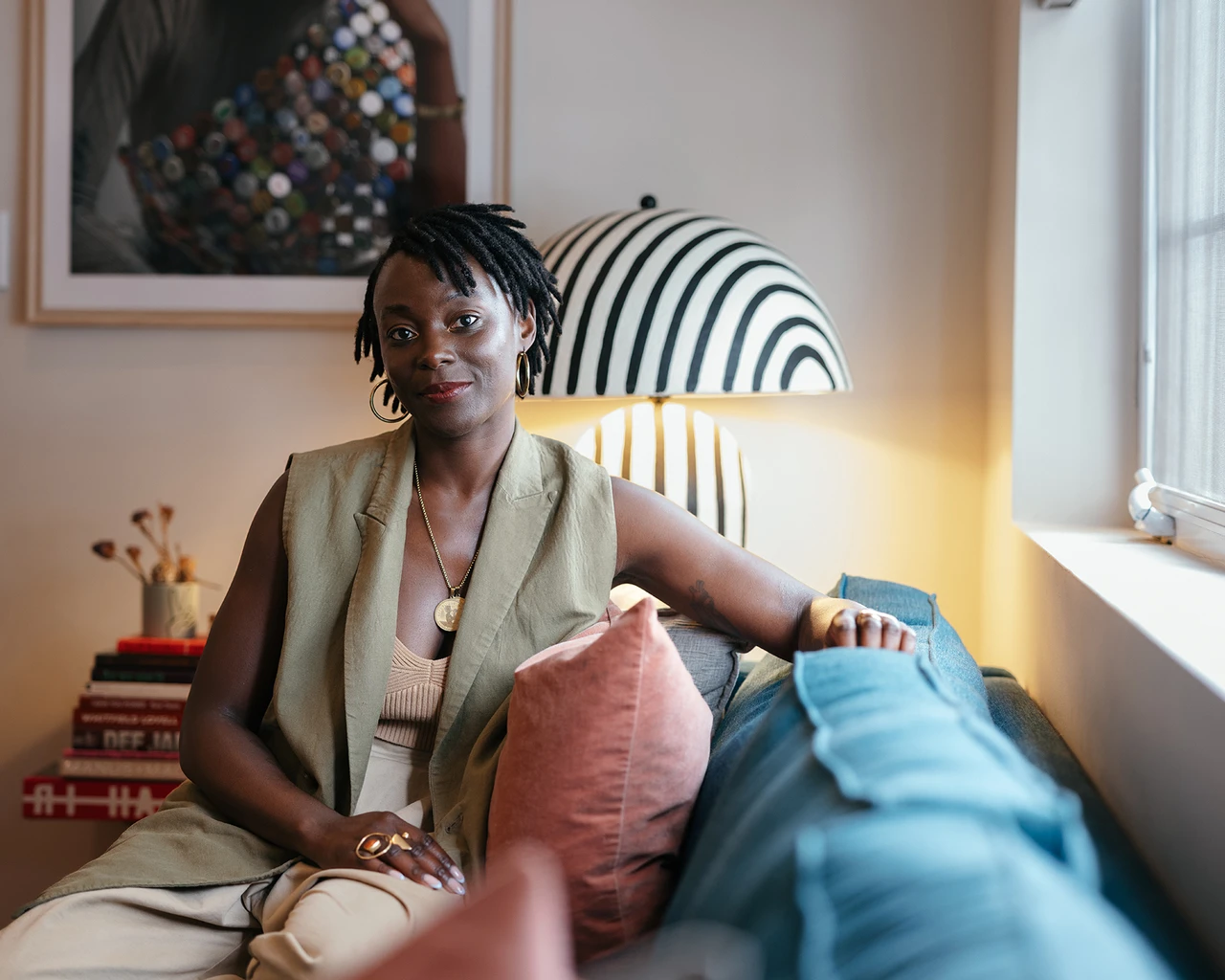 A Black woman in a tan blouse sits on a cushioned couch in front of a framed painting.