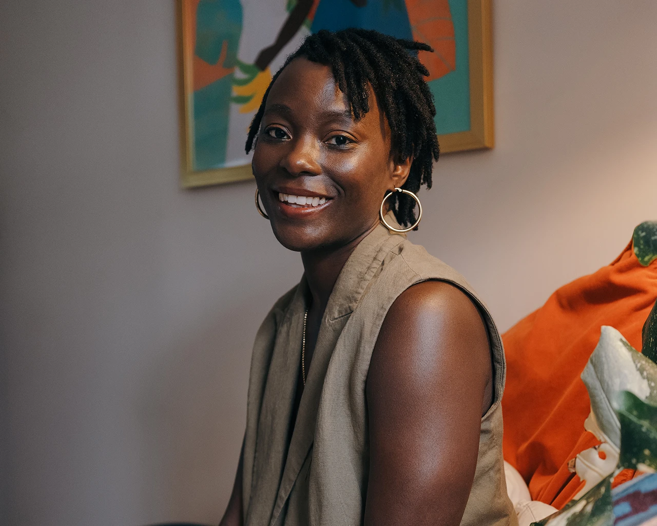 A Black woman in a tan blouse sits on a cushioned couch in front of a framed painting.