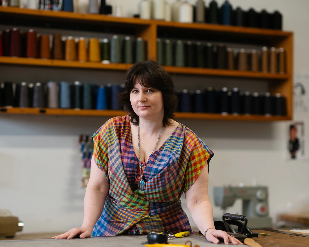 A white woman in a multicolored checkered blouse stands in front of shelving filled with spools of thread.