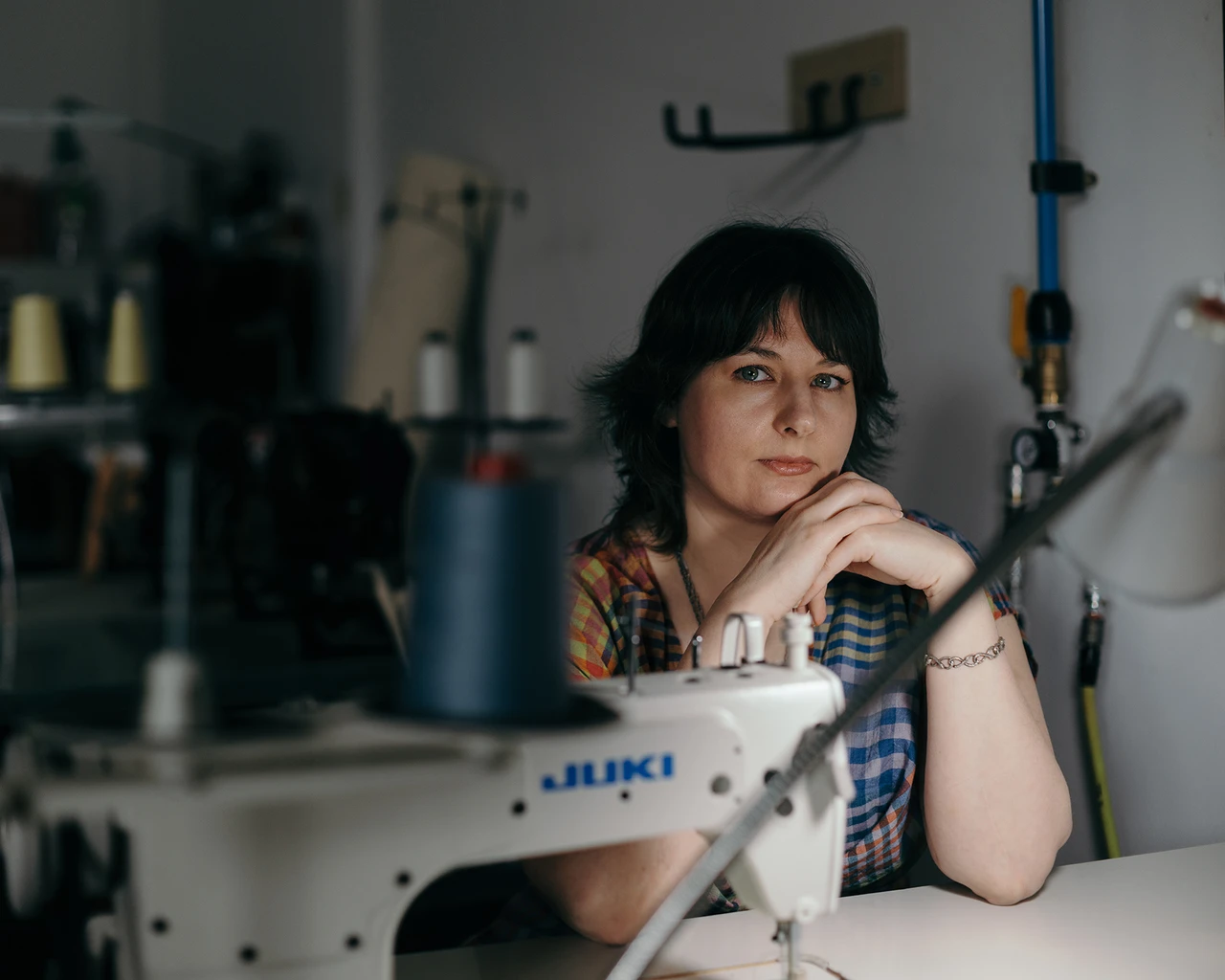 A white woman in a multicolored checkered blouse sits behind a sewing machine.