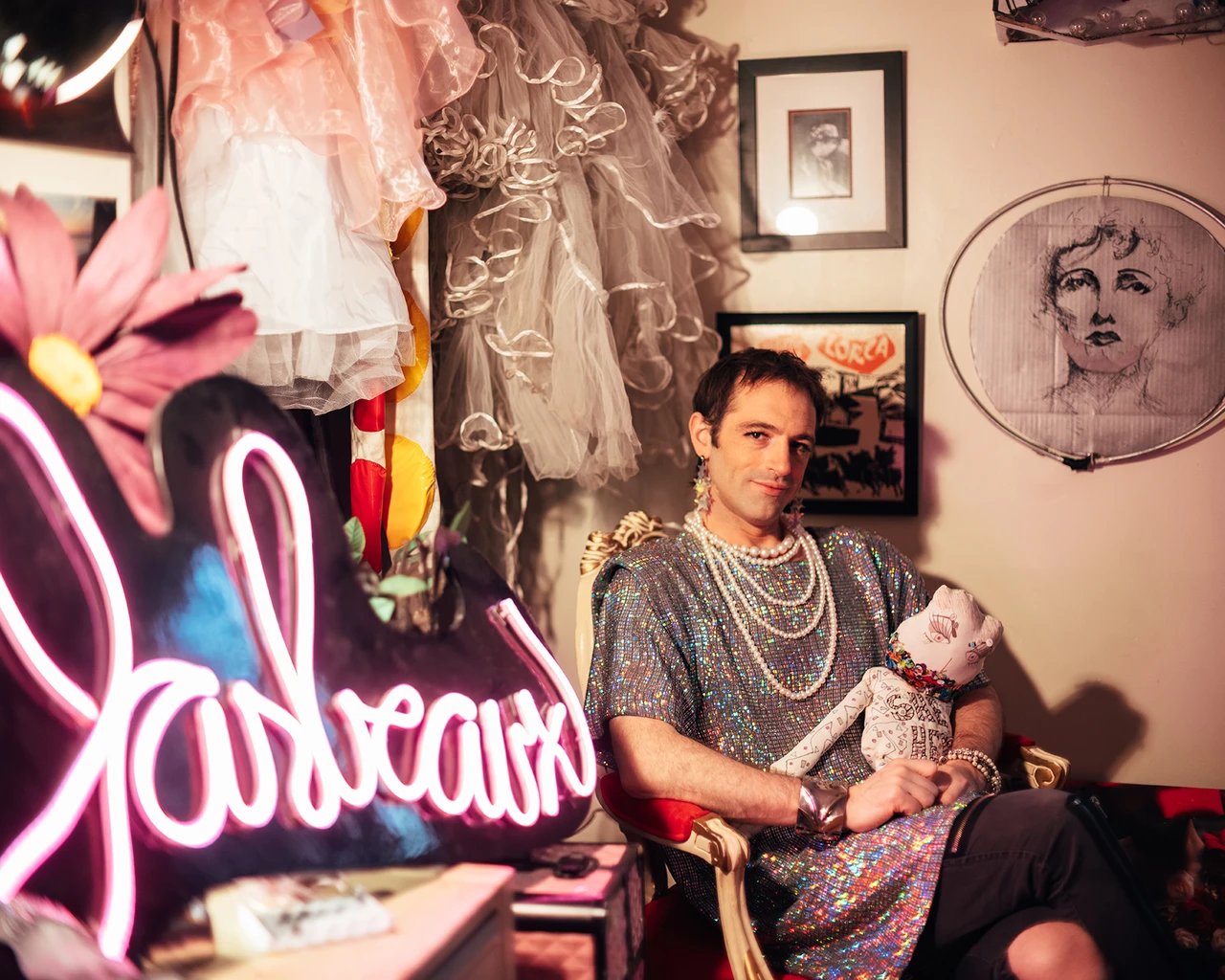 A white woman in a pinkish-grey blouse and costume jewelry sits at a dressing room mirror next to a neon sign.