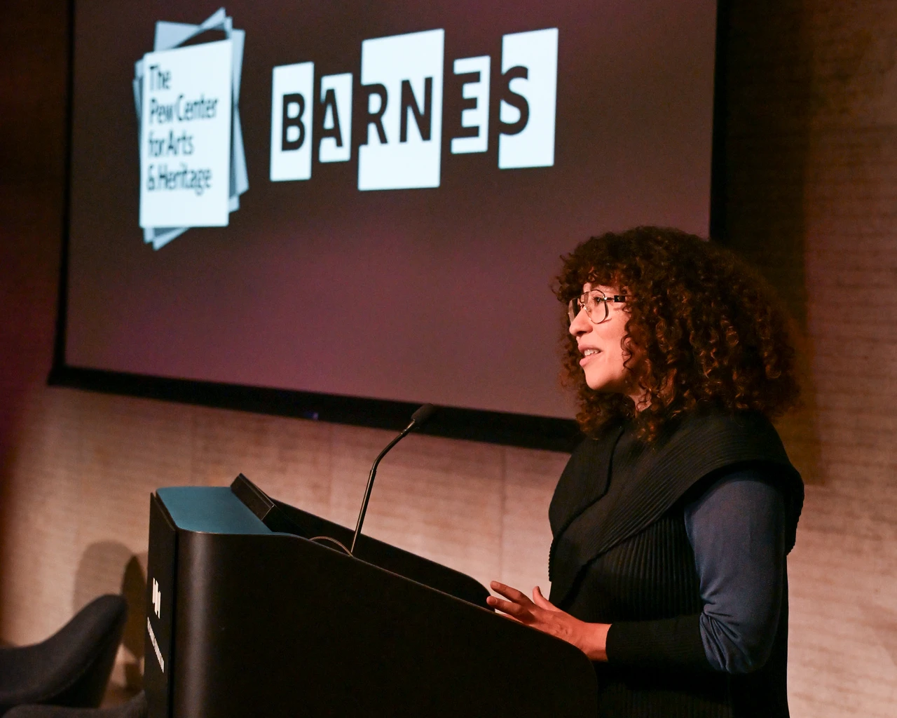 A person stands at a podium speaking in front of a large screen displaying the text "On Practice: Pew Fellows In Conversation," along with logos for "The Pew Center for Arts & Heritage" and "Barnes" as sponsors.