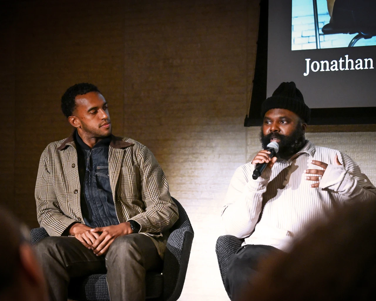 Two people are seated and speaking at an event. One is holding a microphone, wearing a beanie and light-colored top. The other is listening attentively, wearing a striped jacket. A partial screen in the background displays the name "Jonathan González.".