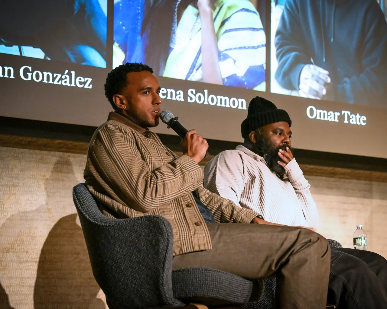 A person is speaking into a microphone, seated next to another individual who has a contemplative expression. They are on a stage with a backdrop displaying names: Jonathan González, Sosena Solomon, and Omar Tate.