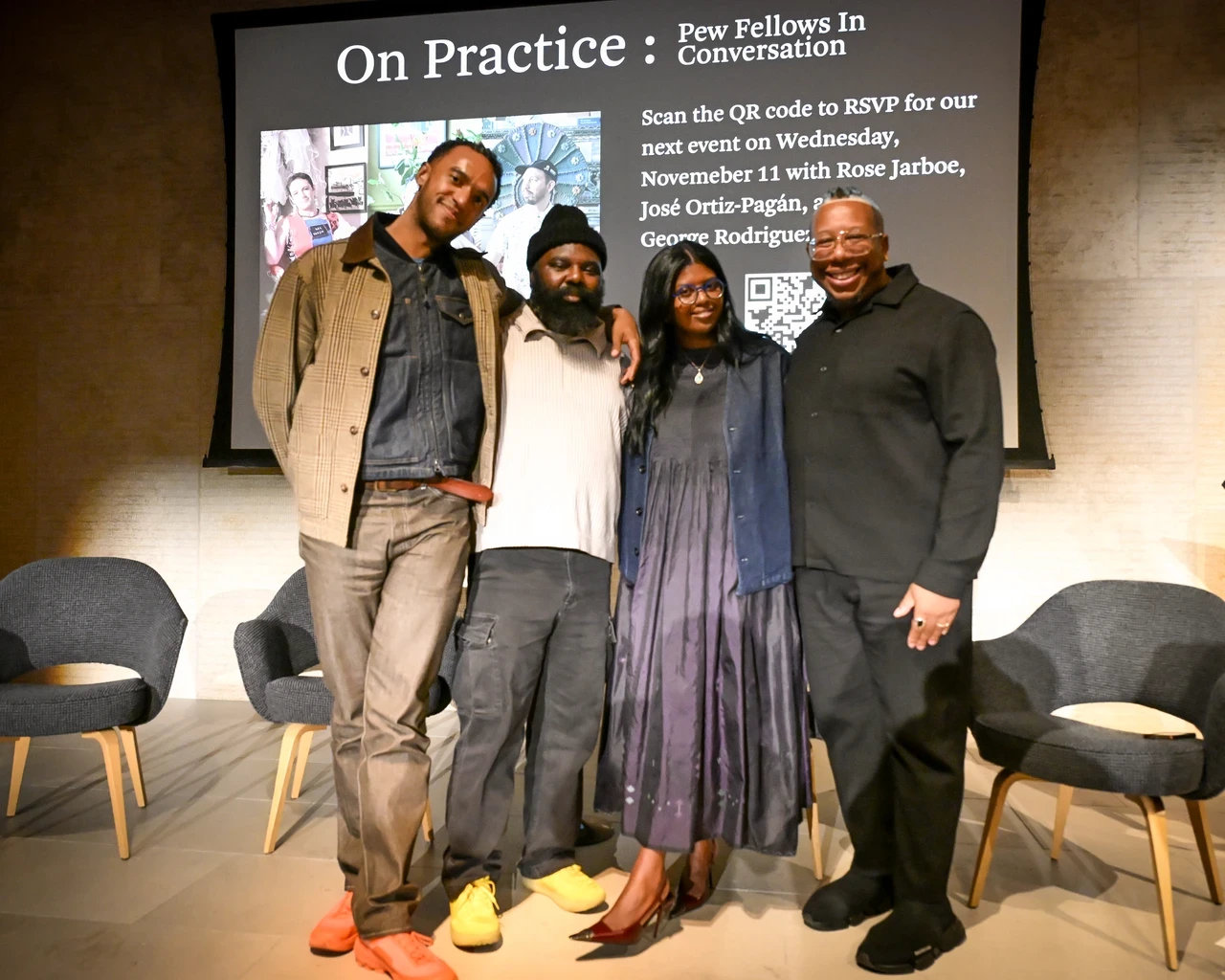 Four people stand together in front of a projected presentation slide titled "On Practice: Pew Fellows In Conversation." They are posing for a photo on a stage with two empty chairs beside them.