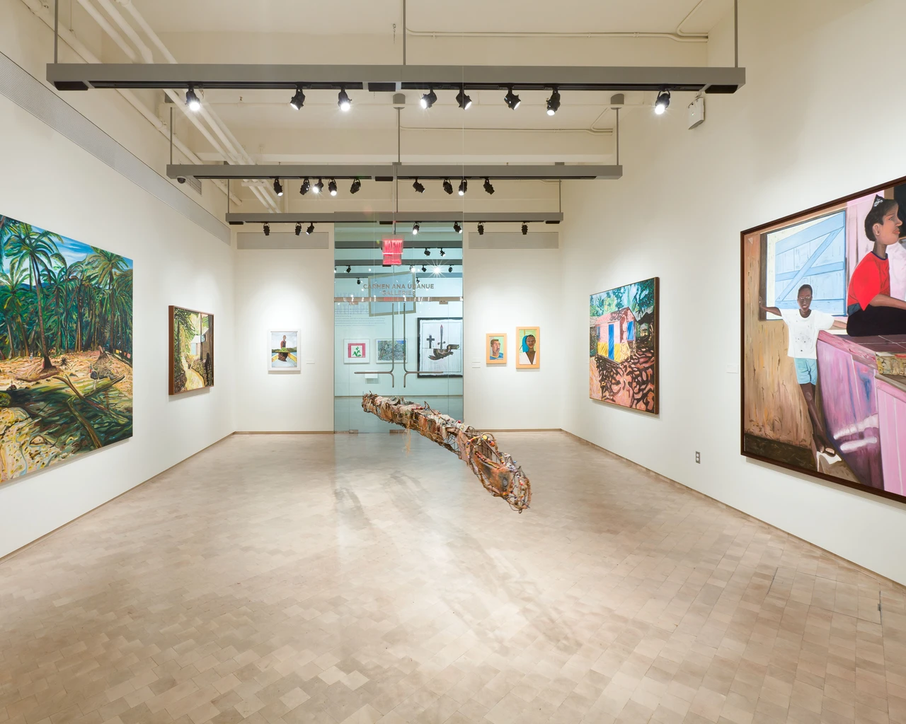 A spacious art gallery featuring various paintings on white walls, with landscapes and portraits. A long, wooden sculpture is displayed in the center of the room. The gallery has overhead track lighting and a glass wall at the far end.