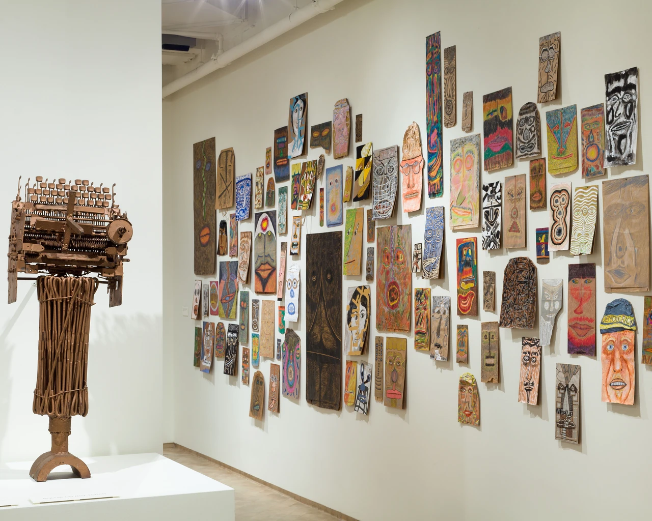 An art gallery with a wooden sculpture on a pedestal to the left and a large, colorful collage of various artworks on the wall to the right. The artworks include abstract designs and faces in vibrant colors. The setting is well-lit and minimalistic.