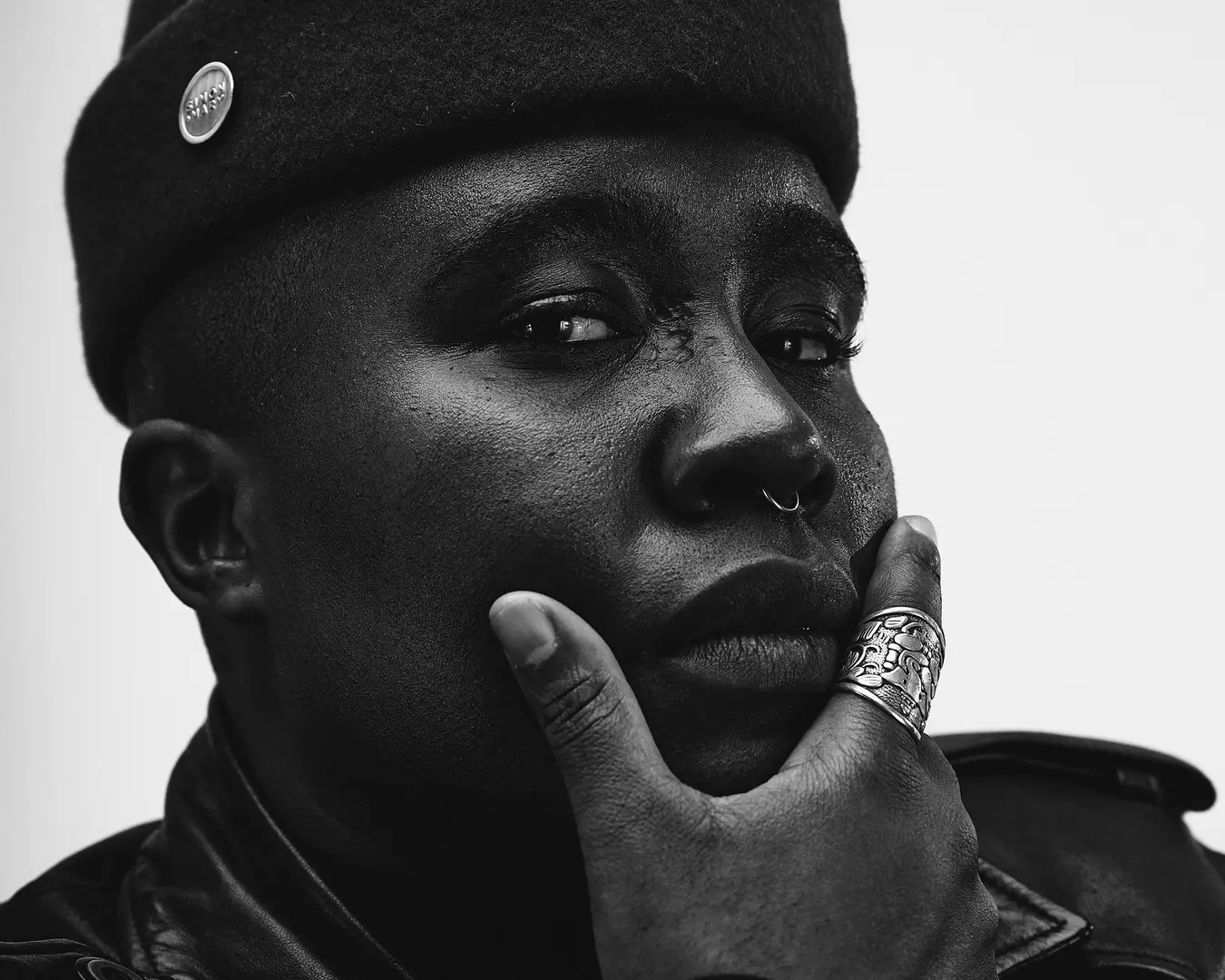 A Black Nonbinary person looks directly at the camera with their right hand cupping their chin. They are wearing a tall cap and a leather jacket with rings. They have a septum piercing and prideful expression. The person's skin and clothing are various shades of black and the background is white.