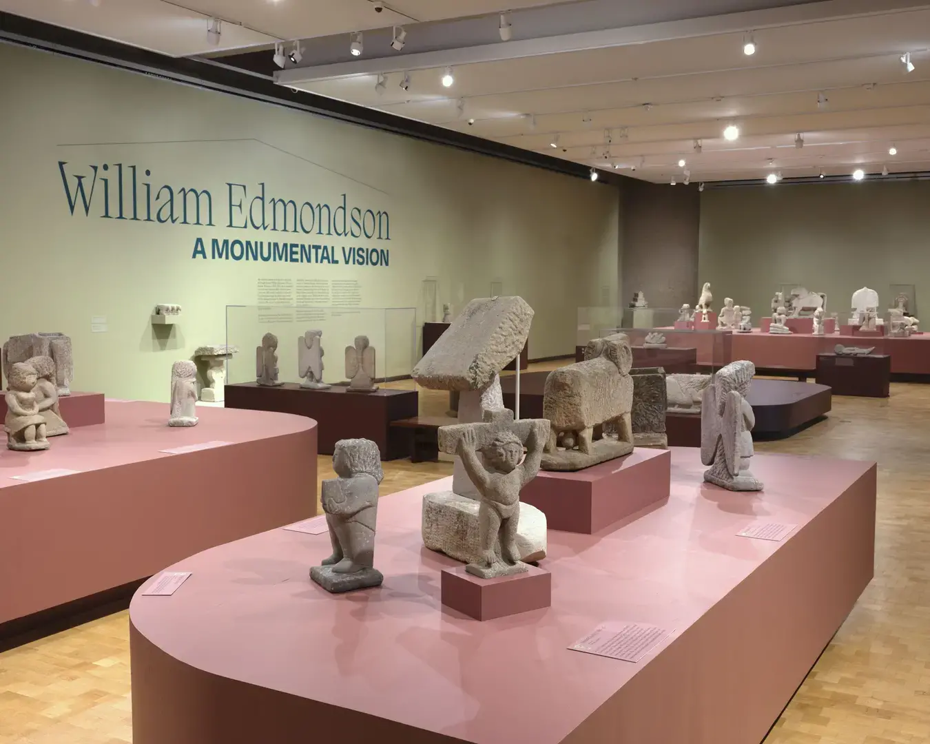 An exhibition of carved stone sculptures on salmon pink pedestals. Painted on the wall in dark green is the exhibition title: "William Edmondson: A Monumental Vision."