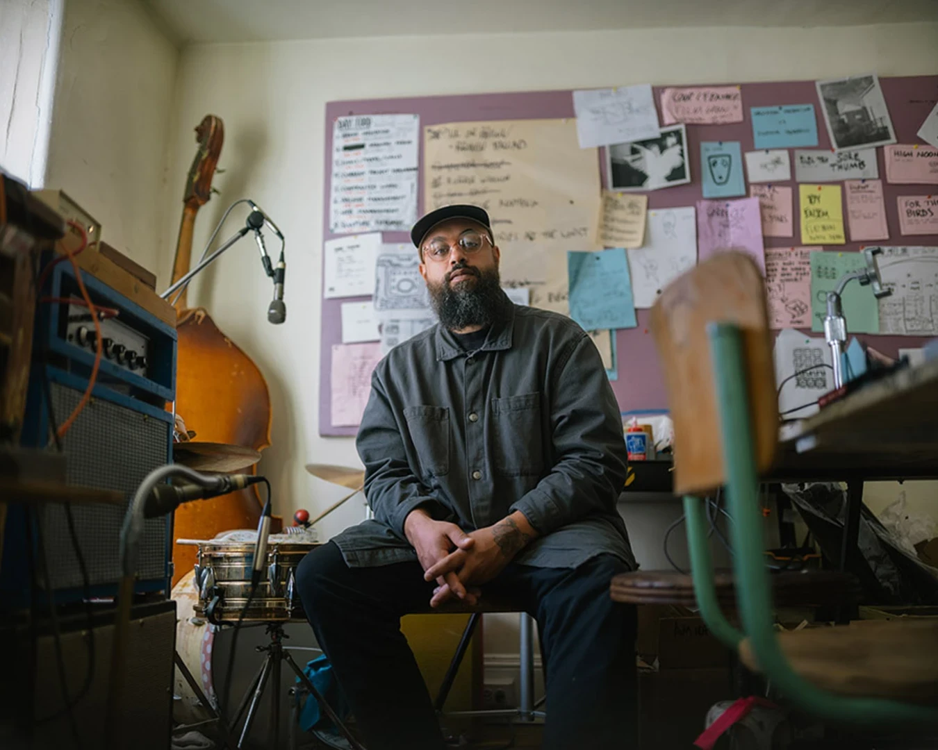A man with a beard wearing glasses, a hat, and a blue shirt sits in a room surrounded by instruments and poster art.