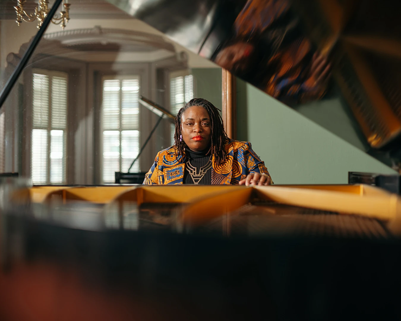 A Black woman in a yellow and maroon blazer sits at a grand piano with the lid opened.