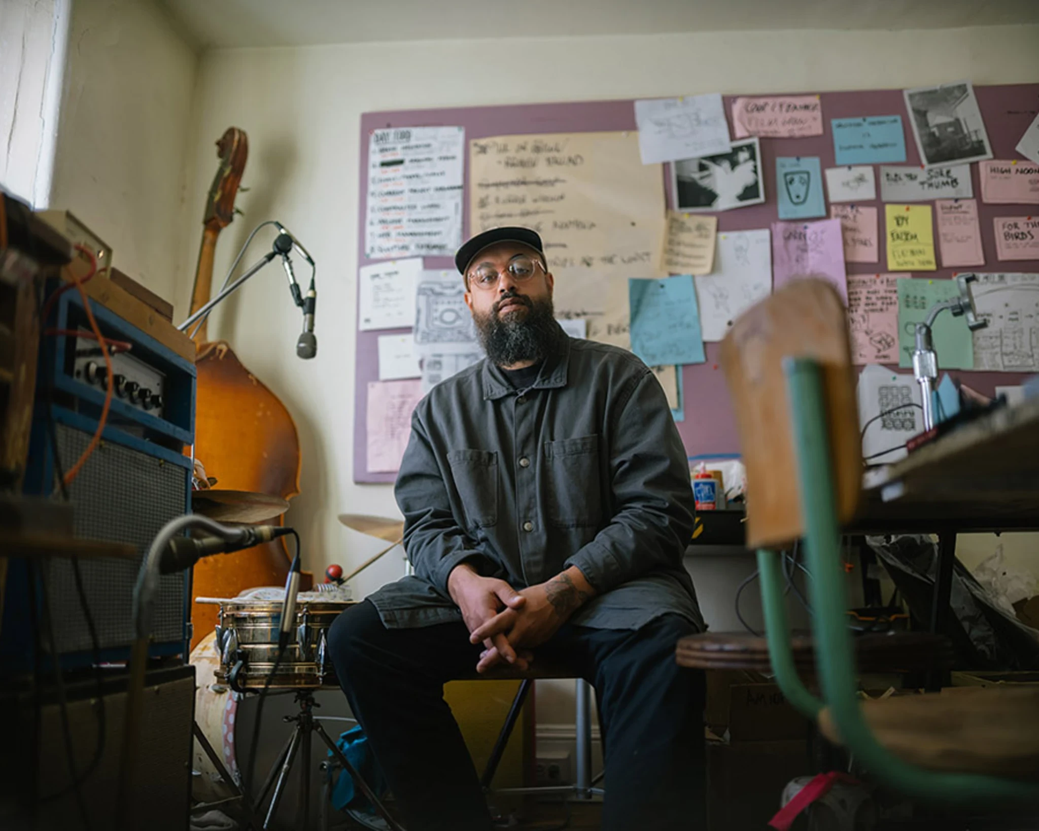 A man with a beard wearing glasses, a hat, and a blue shirt sits in a room surrounded by instruments and poster art.