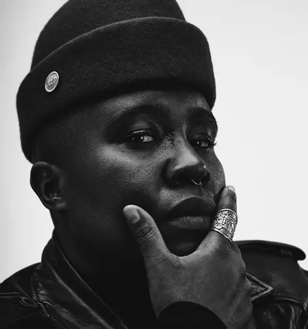 A Black Nonbinary person looks directly at the camera with their right hand cupping their chin. They are wearing a tall cap and a leather jacket with rings. They have a septum piercing and prideful expression. The person's skin and clothing are various shades of black and the background is white.