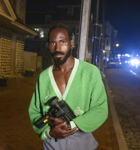 A portrait of filmmaker Vernon Jordan. He stands outside at night holding his video camera. He is a Black man with facial hair wearing a green v-neck cardigan.