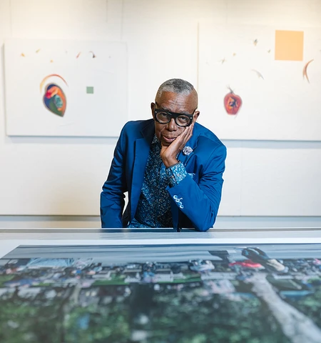 A Black man in a blue blazer and paisley shirt with glasses sits in front of a canvas.