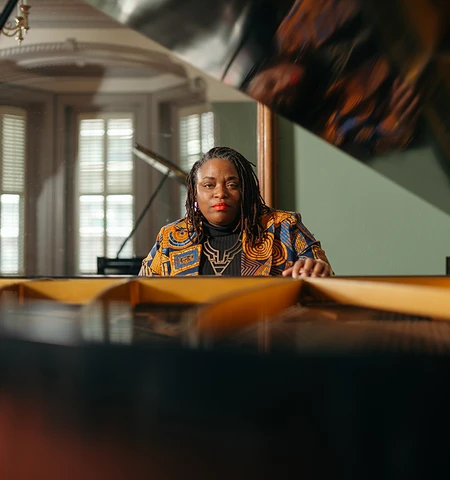 A Black woman in a yellow and maroon blazer sits at a grand piano with the lid opened.