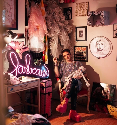 A white woman in a pinkish grey blouse and costume jewelry sits at a dressing room mirror.