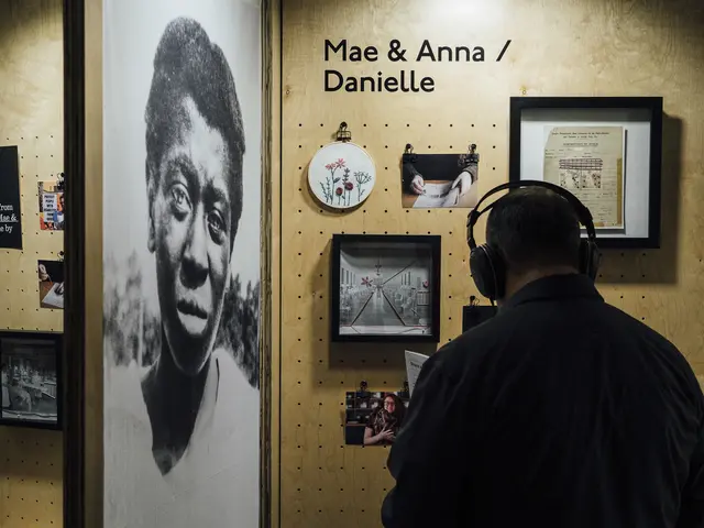 A man wearing headphones stand in front of a wooden display that features framed embroidery, archival imagery and contemporary photos. The words Mae & Anna / Danielle appear on the display. Adjacent to the display is a larger than life-sized archival portrait of a middle-aged Black woman.