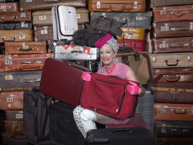 A white woman in a pink flight attendant's dress and hat holding an unzipped suitcase sits in front of a wall of vintage suitcases