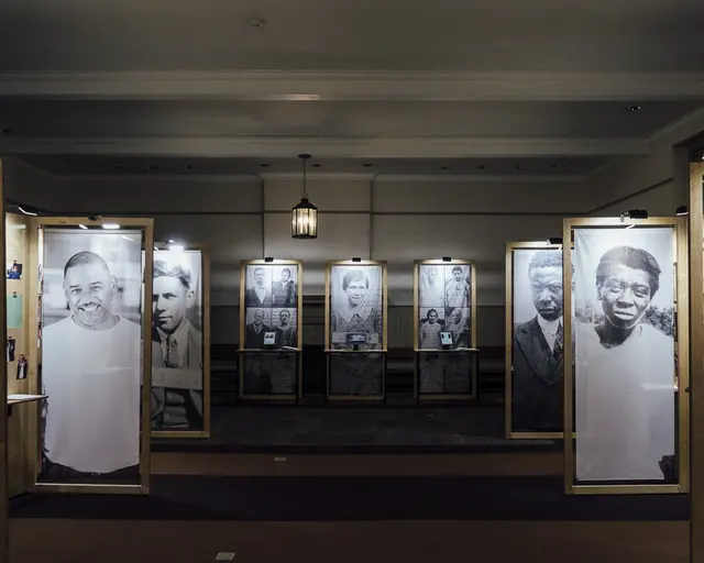 Six, larger than life-sized portraits of men and women line either side of a carpeted room. The images are printed on fabric and hung from wooden frames. In the back of the room, three additional wooden frames; the center frame contains a portrait, and the frames on either side of the portrait contain a grid of six images each and abstracted archival text. All but two of the portraits in the room are archival.