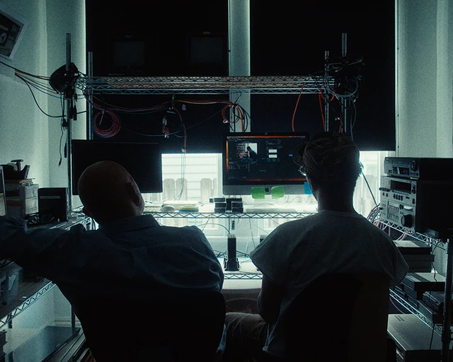 Two men, Zac Manuel and his father Phillip, sit in a dimly-lit video editing station, silhouetted by the greyish-blue light of their computer screen